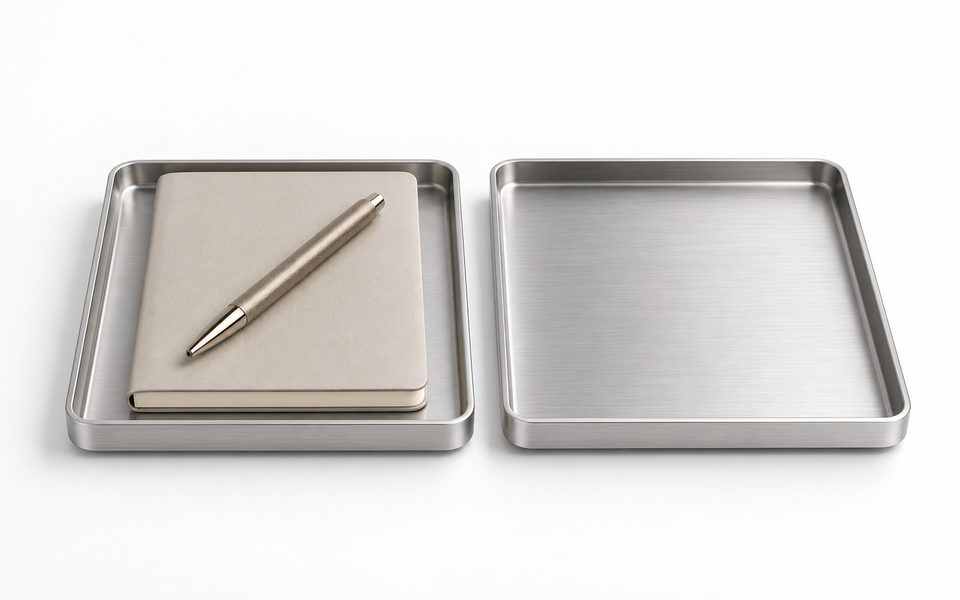 Two brushed-aluminium trays on white, one holding a closed notebook with a machined steel pen laid diagonally across it, the other empty.