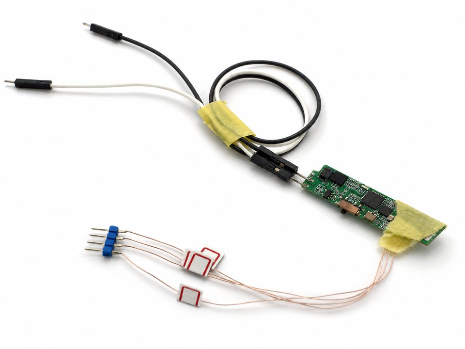 A small green controller board wired by hand to two piezo sensor tiles, taped at the cable break, shown against a clean white ground.
