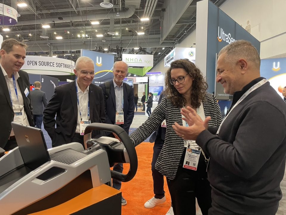 A trade-show stand with visitors gathered around a steering-wheel demonstration and a display screen behind it.