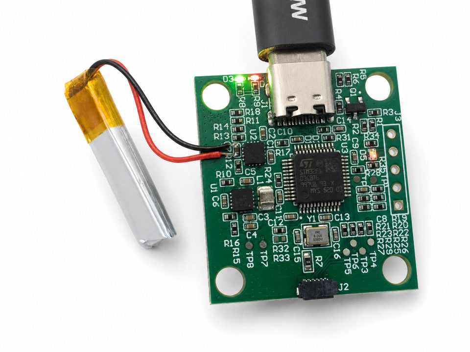 A green controller bring-up board with a hand-soldered battery pack taped alongside it and a USB-C connector, on a clean white ground.