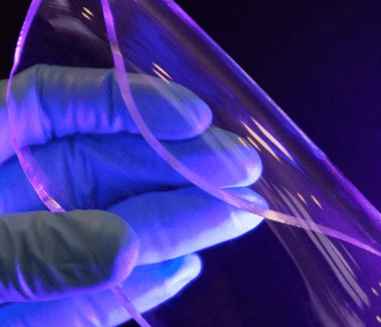 Gloved fingers bending a thin transparent piezoelectric film, lit from behind in violet so the curve of the film catches the light.