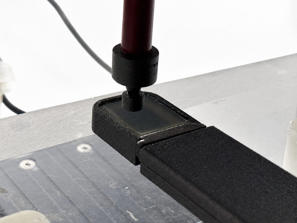 A force-test rig -- a cylindrical probe on a vertical post pressed down onto a black test coupon clamped to a metal base plate.