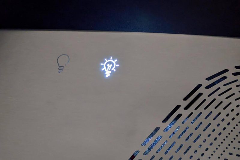 A brushed metal panel with a single small blue-white icon lit through the metal, and a row of fine ventilation perforations beside it.