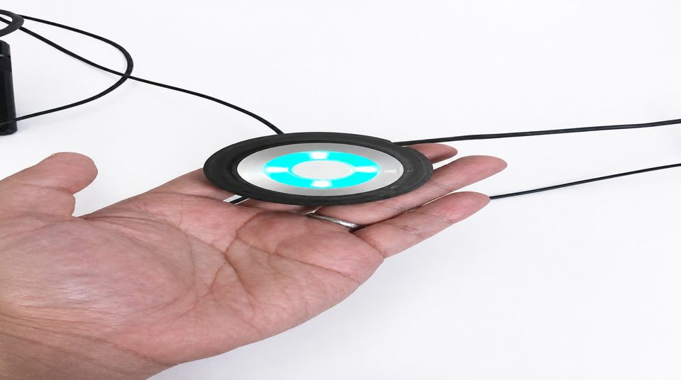An open hand holding a small round black sensor module with a teal ring lit around its face.