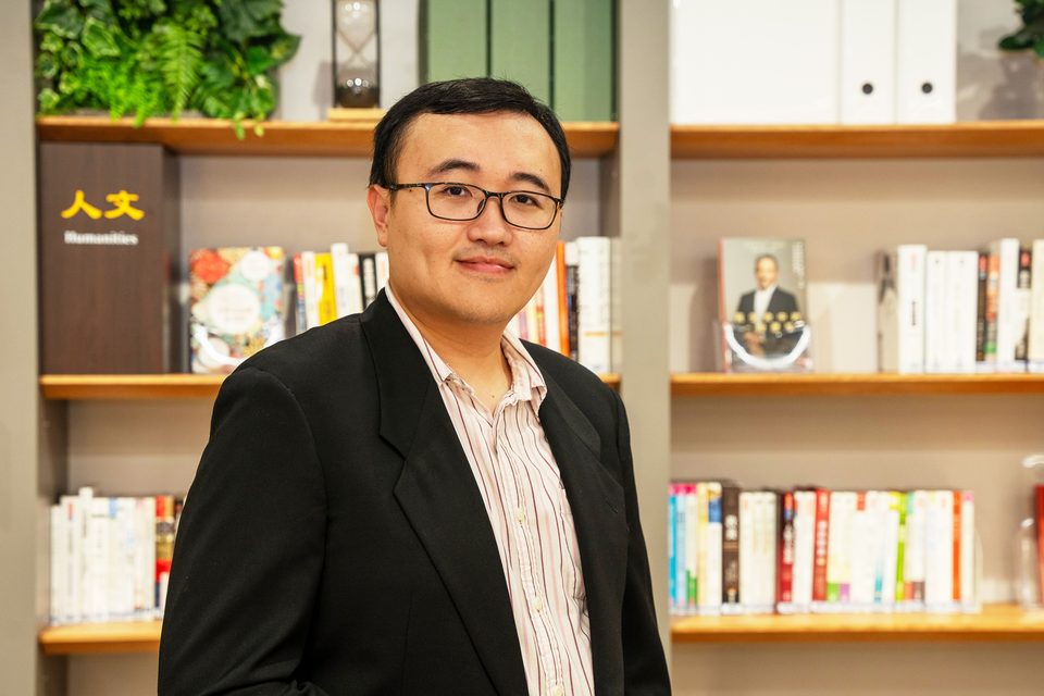 Hao-Yen Tang in a dark jacket, photographed standing in front of a library shelf of books and files.