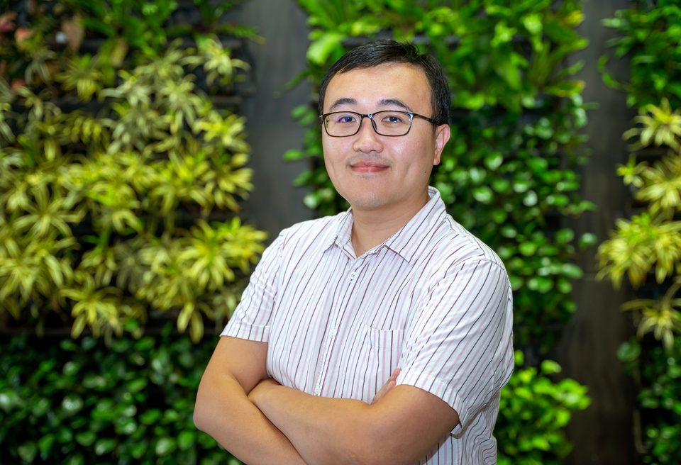 Hao-Yen Tang in a striped shirt with his arms folded, photographed smiling against a wall of green planting.