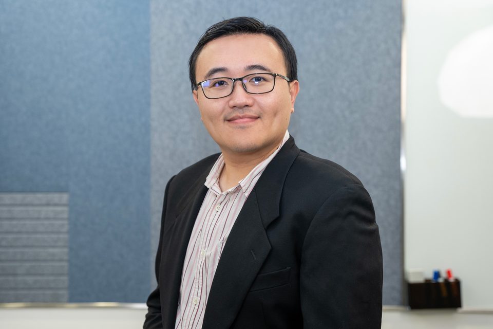Hao-Yen Tang in a dark jacket and striped shirt, turned three-quarters to camera, photographed against a grey office wall.