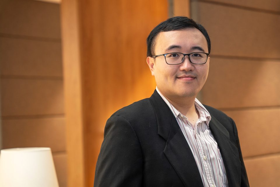 Hao-Yen Tang in a dark jacket, photographed in warm light against a wood-panelled wall with a lamp out of focus beside him.