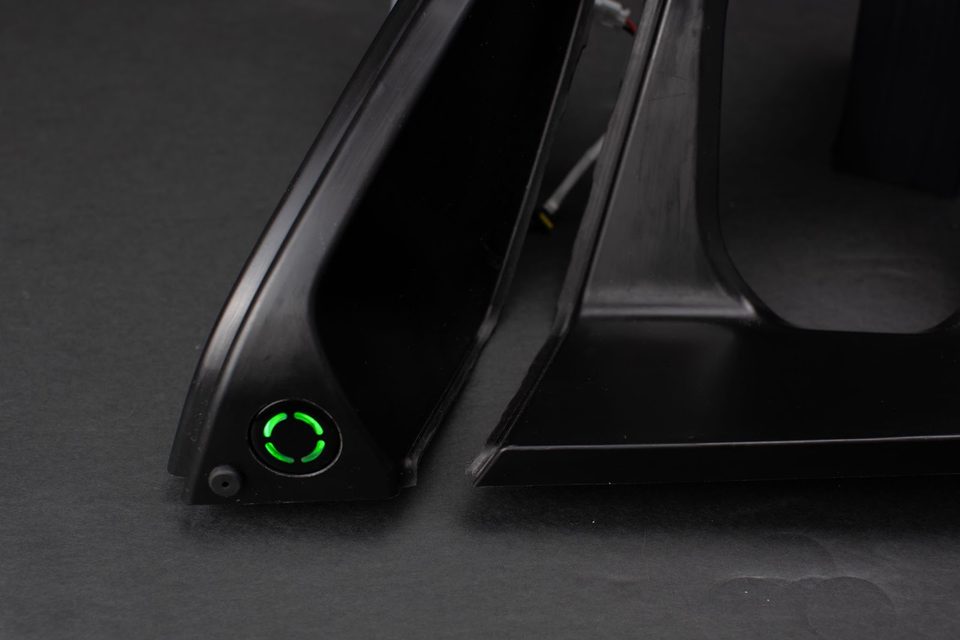 A dark automotive steering wheel photographed close, with a small illuminated green ring glowing through the solid trim where a control sits.